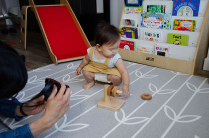 Curious baby girl attentively examining the third wooden ring for the Cubos Horizontal Dowel Rings Stacker, observing its size and shape, and contemplating her next move in this engaging and educational playtime activity. Her inquisitive nature is evident as she explores and learns through hands-on play.