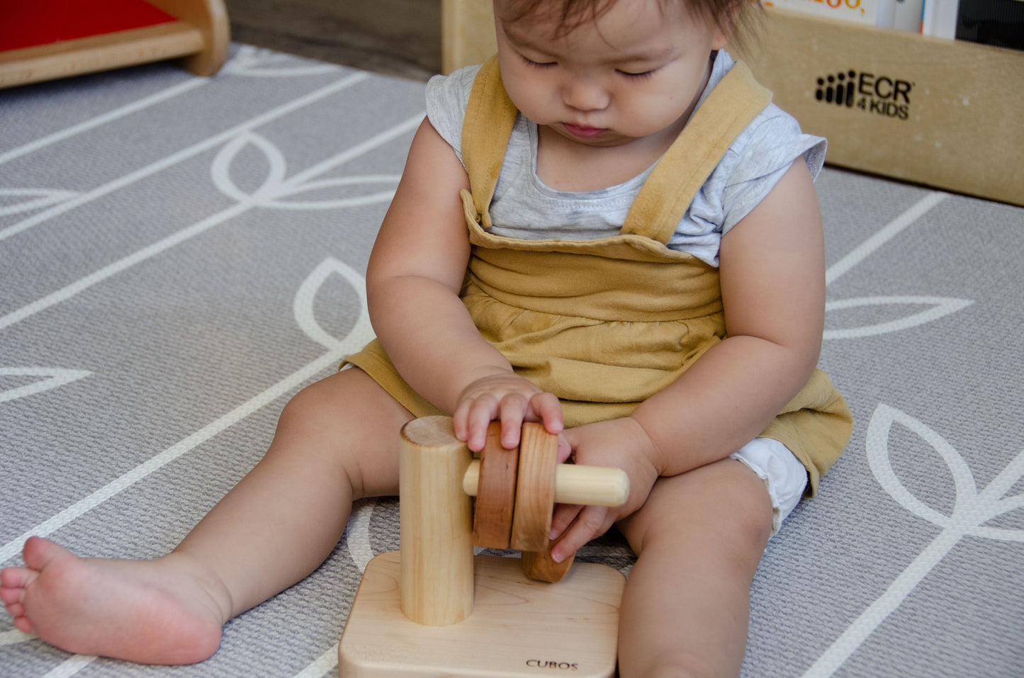 Adorable baby girl attentively placing the wooden ring on the Cubos Horizontal Dowel Rings Stacker, focused on the task and learning the concept of stacking, all while having a fun and rewarding playtime experience.