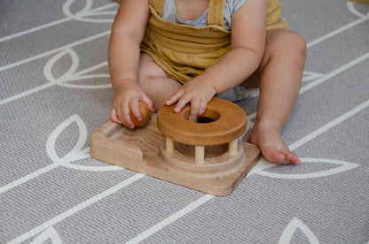 The baby girl is engrossed in play with the Permanence Box, a classic Montessori toy. With great curiosity and excitement, she explores the various compartments, practicing her hand-eye coordination and fine motor skills as she discovers the joy of fitting objects into their designated spaces. This educational and interactive playtime experience helps her develop an understanding of object permanence and spatial relationships, providing a foundation for her cognitive development.